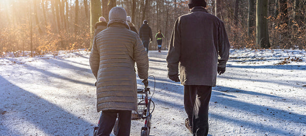 Elderly couple walking along a snowy woodland path in winter, with one person using a rollator walker as others walk ahead in the distance. Image