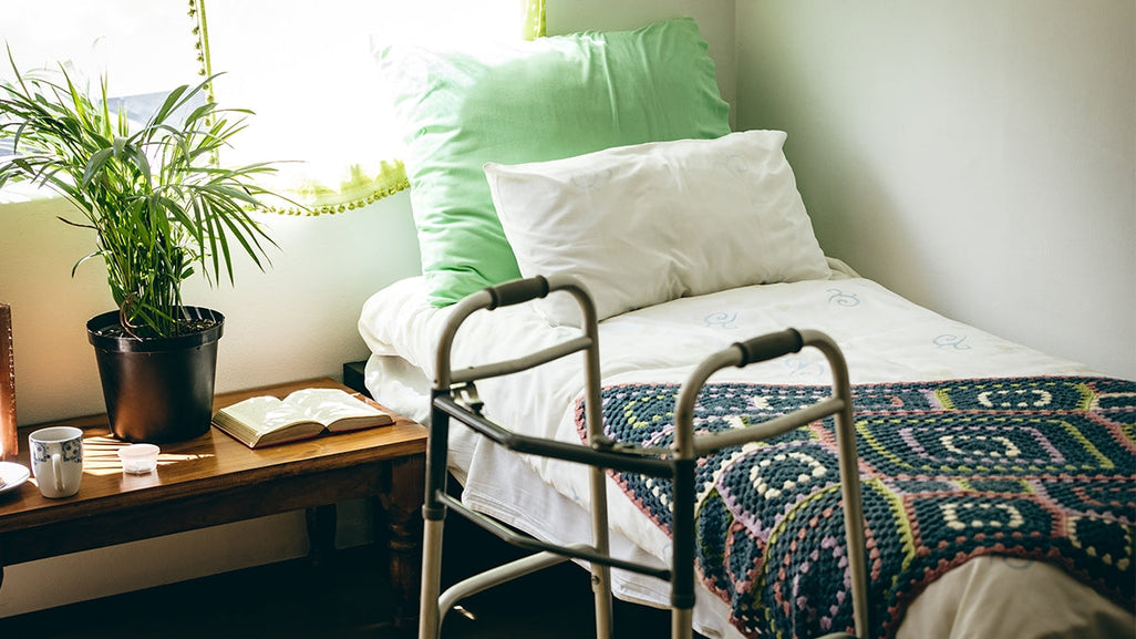 A cozy bedroom with a crochet blanket on the bed, green pillows, a wooden table with a plant and book, and a walker by the window.