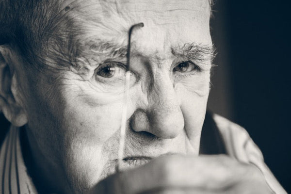 An elderly man thoughtfully holds a thin object near his face, wearing a striped shirt; background is softly blurred.