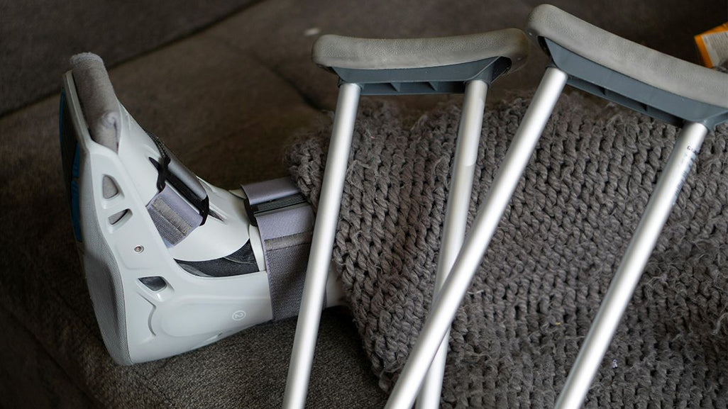 A leg in a medical boot is elevated on a gray sofa with a chunky blanket. Two gray crutches lean against the sofa.
