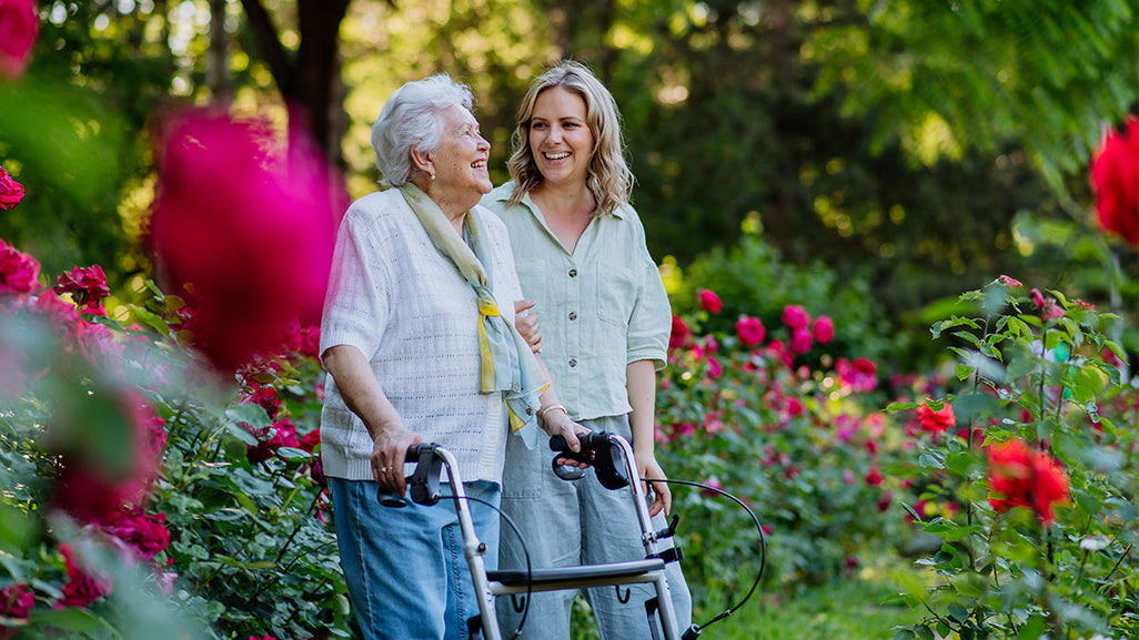 An elderly woman with a walker and a smiling younger woman walk through a rose garden, surrounded by red roses and sunlight filtering through trees.