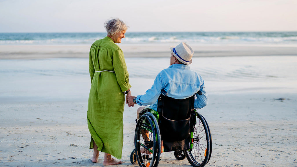An elderly couple on a beach. The woman in a green dress holds hands with a man in a wheelchair wearing a hat and blue shirt.