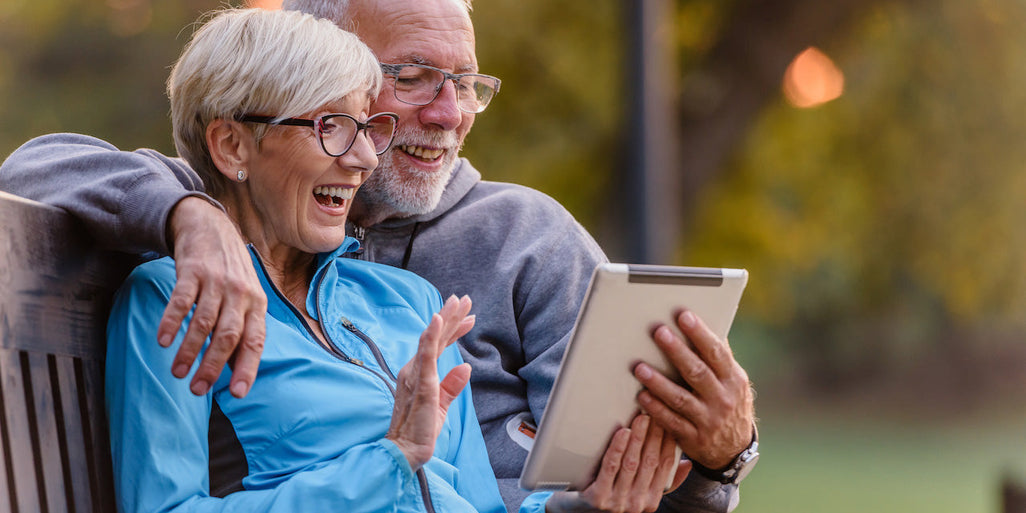 An elderly couple sits on a bench, smiling at a tablet. The woman wears a blue jacket; the man, a gray hoodie. Greenery surrounds them.