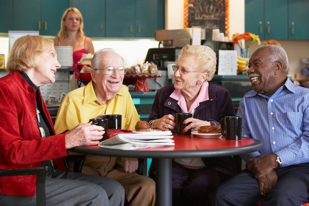 Four elderly people sit at a cafe table, smiling with coffee cups. A woman stands by baked goods counter. Warm, inviting atmosphere.