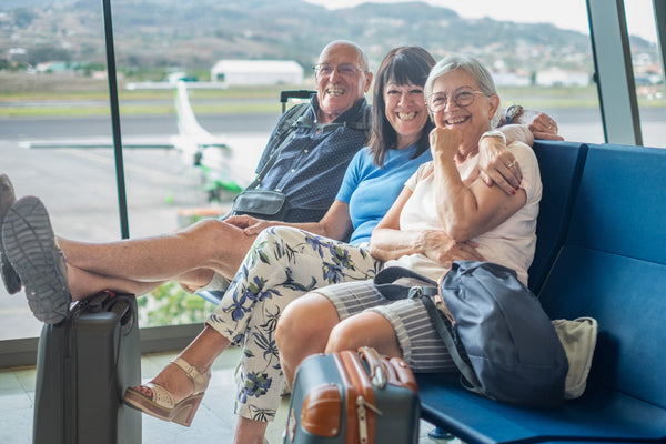 Three people sit on blue airport chairs. Two relax with feet on luggage, one in blue shirt. Large windows show an airport runway and plane.
