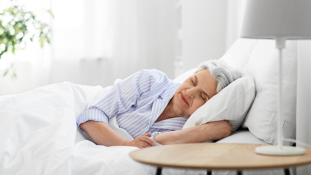 An older adult sleeps peacefully in bed, wearing light blue striped pajamas. A plant and wooden table are visible in the softly lit room.