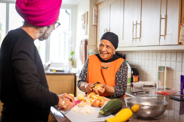 An elderly woman in an orange apron peels vegetables, smiling. A man in a pink turban slices food nearby amidst kitchen tools and ingredients. Image