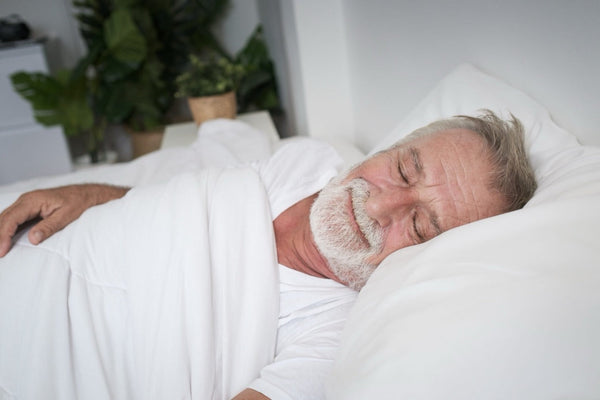 An elderly man sleeps peacefully on a bed with white sheets; a potted plant sits near the small dresser in the softly lit room.