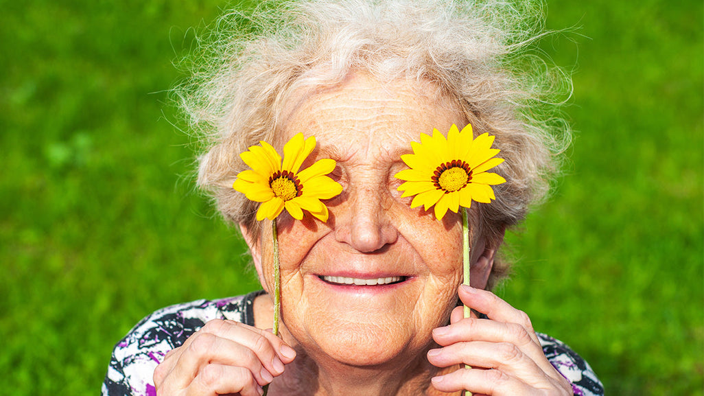 An elderly person joyfully holds two yellow flowers over their eyes, smiling against a bright green background.