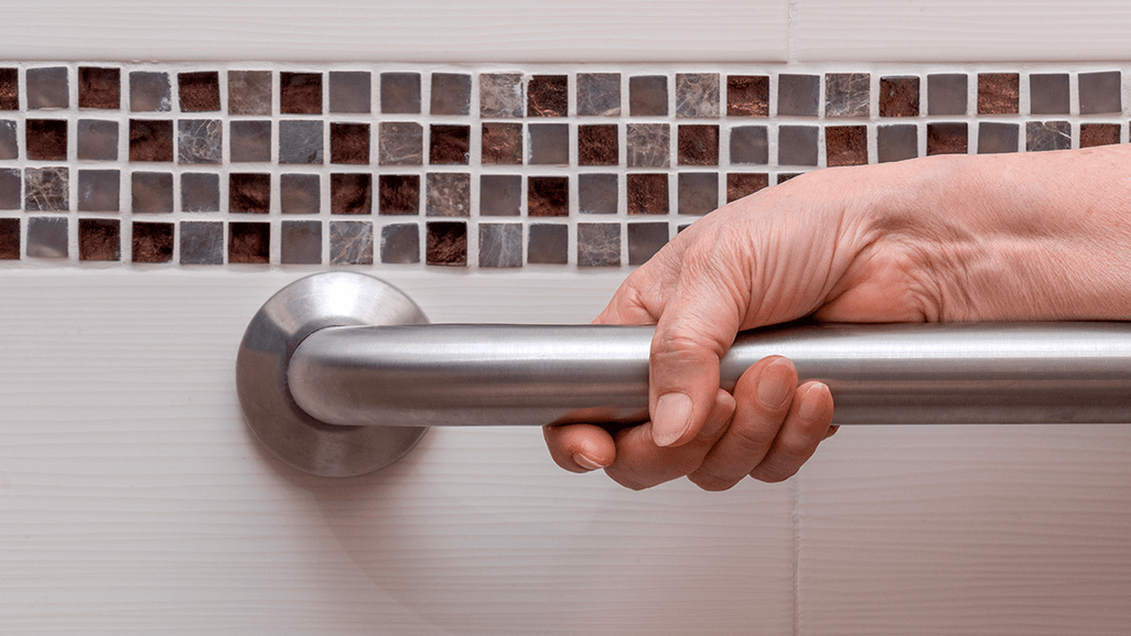A hand holds a metal grab bar on a white tiled bathroom wall with a brown and gray mosaic strip, highlighting bathroom safety.
