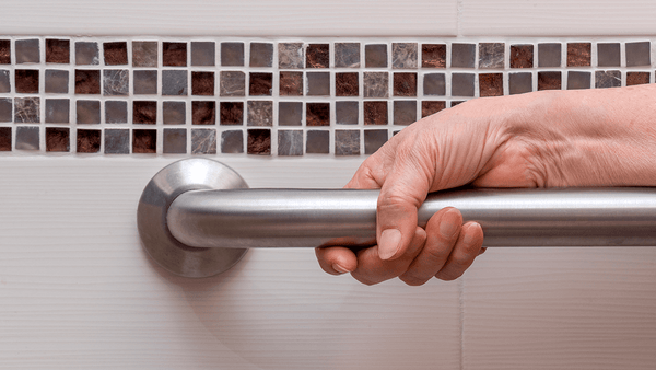 A hand holds a metal grab bar on a white tiled bathroom wall with a brown and gray mosaic strip, highlighting bathroom safety. Image