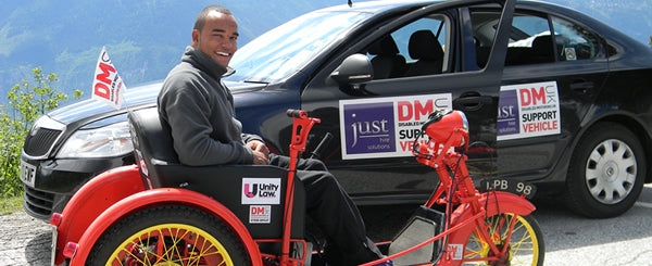 A person sits in a bright red tricycle with yellow wheels beside a black car labeled DM Support Vehicle against a mountain backdrop.