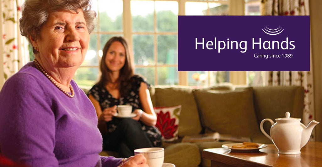 An older woman in a purple sweater enjoys tea at a warm table, smiling. A younger woman also drinks tea behind her.
