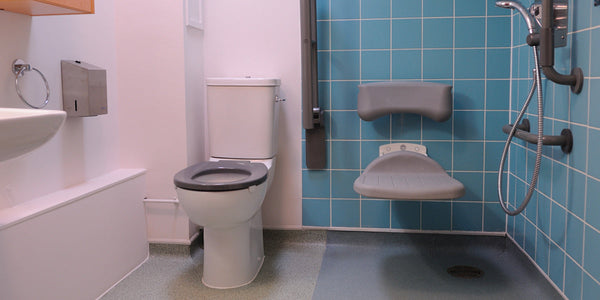 A bathroom with a white toilet, gray grab bars, a wall-mounted shower; light blue tiled shower area with a foldable gray seat.