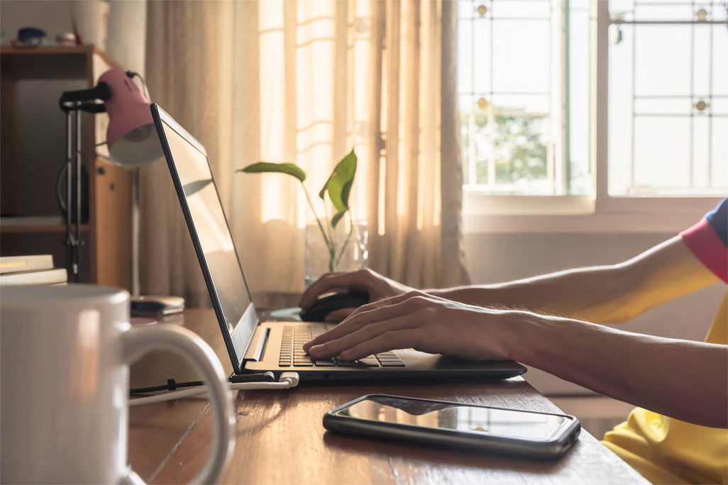 A person types on a laptop at a wooden desk by a sunlit window with curtains. A phone, coffee mug, and plant are on the desk.