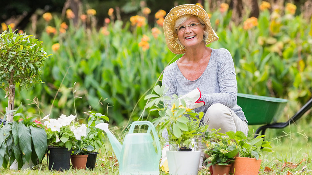 A woman kneels in a garden with potted plants, holding gloves near a watering can, surrounded by tall orange flowers and green leaves.