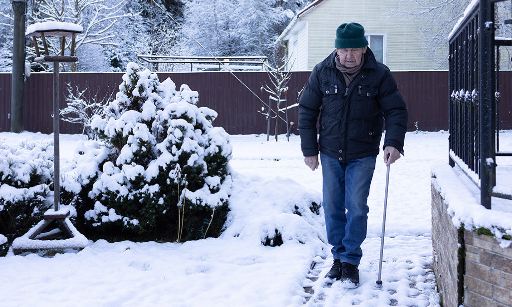Older man walking carefully along a snowy garden path using a walking stick, with winter conditions around his home.