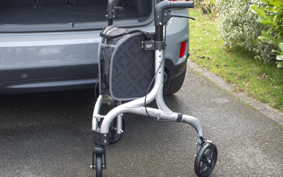 A silver walker with three wheels and a black storage bag stands on a driveway next to an open gray car trunk, with bushes behind it.
