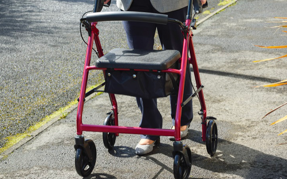 A person using a red walker moves along a sidewalk. They wear dark pants and shoes. The walker has a seat and black handles, with grass nearby.