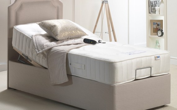 An adjustable bed with a beige headboard, gold pillow, remote, floor lamp, and small shelving unit in the room.