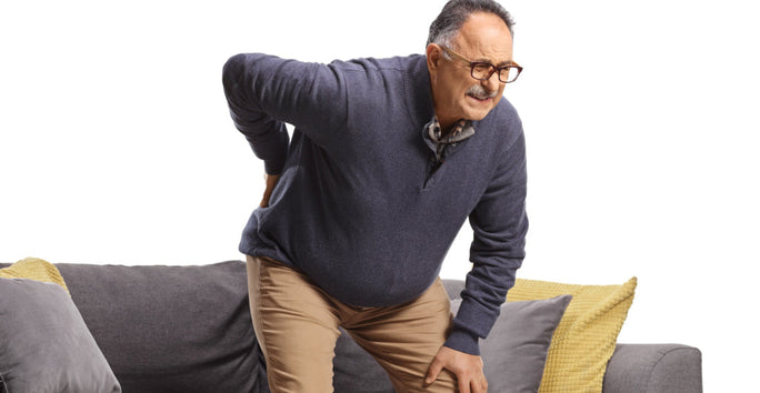 A man in glasses stands up from a couch, holding his lower back with a pained expression. The couch has yellow and gray cushions.