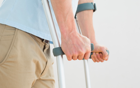 A person in a light blue shirt and khaki pants is holding crutches, focusing on their hands for support. The background is plain white.