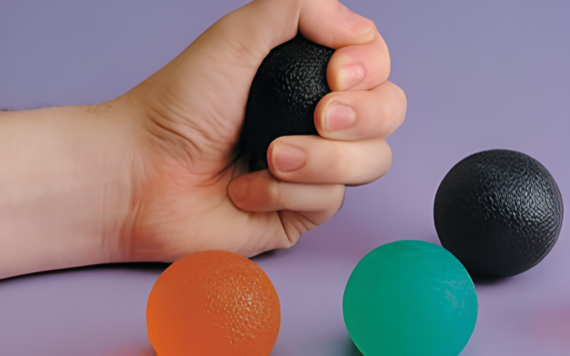 A hand holding a black stress ball with three others—orange, green, and black—on a purple surface and background.