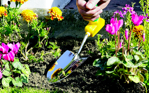A person digs in a garden with a yellow trowel, surrounded by pink cyclamen and orange marigolds under a sunny sky. Image
