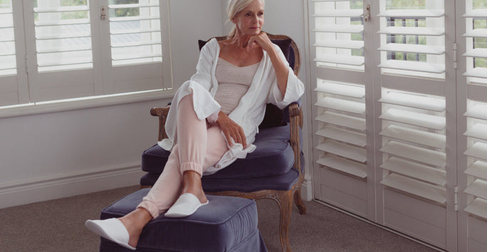 A woman sits on a blue chair in a white robe, light pink pants, and slippers, appearing thoughtful. The room has white shuttered windows.