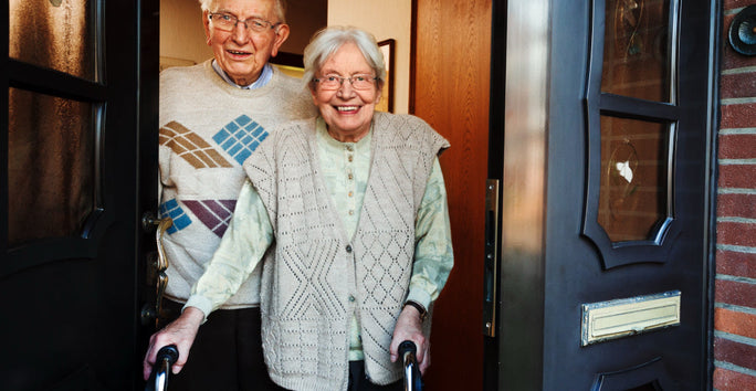 An elderly couple stands at an open doorway. The man, wearing glasses, is beside a smiling woman using a walker. Both are dressed in cozy sweaters, with a warm, welcoming expression on their faces. The door reveals a glimpse of a brick exterior.