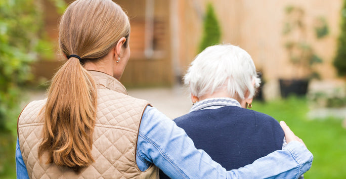 A woman in a quilted jacket helps an elderly person walk in a garden. The scene is warm and caring, with greenery and a blurred building.