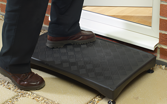 A person steps onto a black textured mobility ramp at a homes entrance, wearing black pants and brown shoes, near a white door threshold.