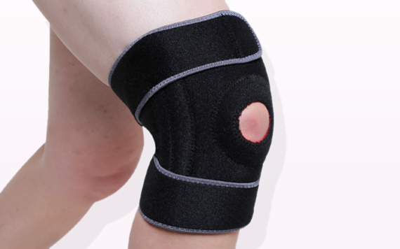 Close-up of a persons knee wearing a black knee brace with straps, showing a circular opening at the kneecap. Background is light pink.