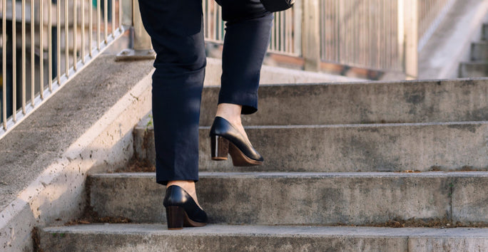 A person wearing black heeled shoes and dark pants is ascending a set of concrete stairs. A metal railing runs alongside the stairs.