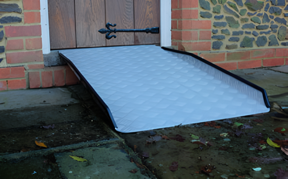 A garden ramp leads up to a wooden door. The ramp has a textured, light gray surface with dark edges. It is surrounded by a mix of brick and stone wall and is placed over a wet, leaf-strewn area.