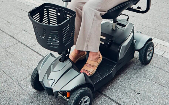 Person sitting on a mobility scooter on a tiled pavement. The scooter has a black basket at the front. The person is wearing beige pants and tan sandals. Only the lower half of the persons body is visible. Image