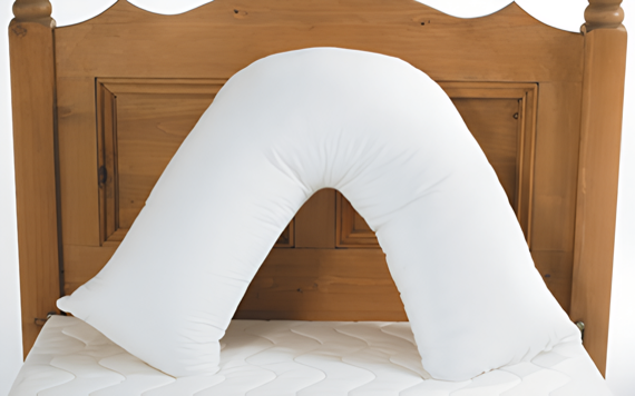 A U-shaped white pillow rests against a wooden headboard on a bed with a white cover, designed to support the back or neck.