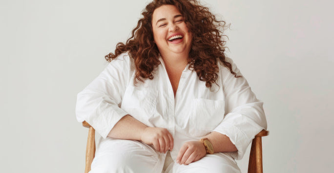 A person is happily sitting on a wooden chair, wearing a white outfit, in front of a light-colored wall.