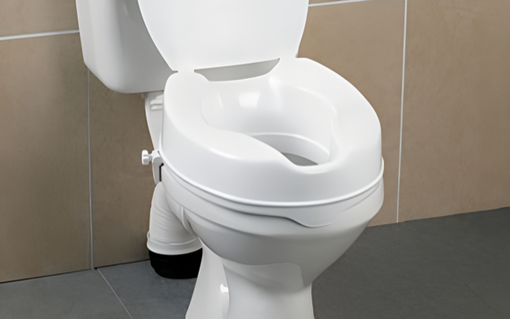 A white elevated toilet seat is on a regular toilet in a bathroom with beige tiles and a gray floor.