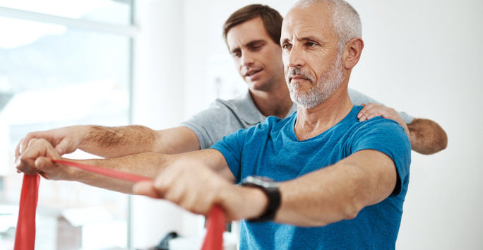 An older man uses a resistance band for exercise, assisted by another man in a well-lit gym or therapy room. Both concentrate on the task.