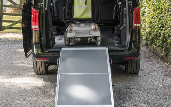 A wheelchair is on a ramp into a black van with open rear doors. The setting is outdoors with a gravel path and greenery.