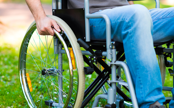 A person in a wheelchair outdoors on grass, gripping the wheel. Sunlight creates shadows; wheels have orange reflectors. Image