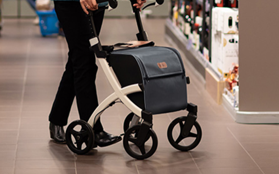 A person uses a rollator with large wheels and a storage compartment in a store aisle, with shelves of products in the background. Image