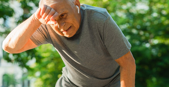 An elderly man stands outside, wearing earbuds and a gray T-shirt. He wipes his forehead, looking tired, against a background of green trees.