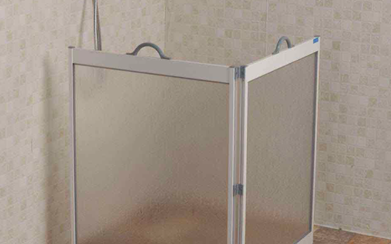 A compact, foldable shower screen with translucent panels, silver frame, and a handle is set up in a bathroom with beige, tiled walls and a wooden floor.