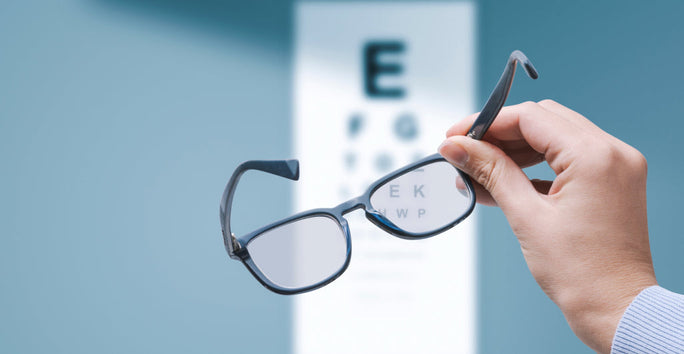 A hand holding eyeglasses in front of a blurred eye chart with letters, against a blue background.