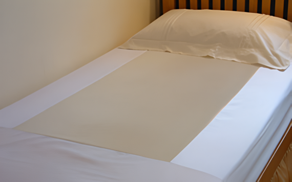 A neatly made bed with white sheets, a beige blanket, and matching beige pillow against a light wall with a wooden headboard.