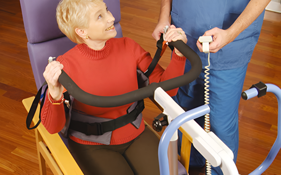 An elderly woman in a red sweater sits smiling in a lift chair. A person in blue scrubs operates it in a room with wooden floors.