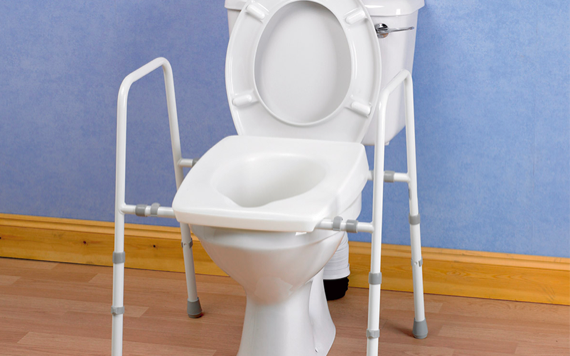 A white toilet with a raised seat has metal safety rails on both sides. It sits on a light brown wood floor against a blue wall.