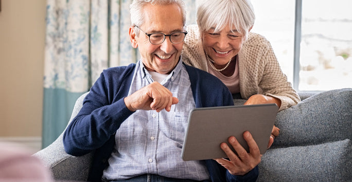An older couple smiles at a tablet, man in glasses on couch, woman leans over. They look happy in a cozy living room.